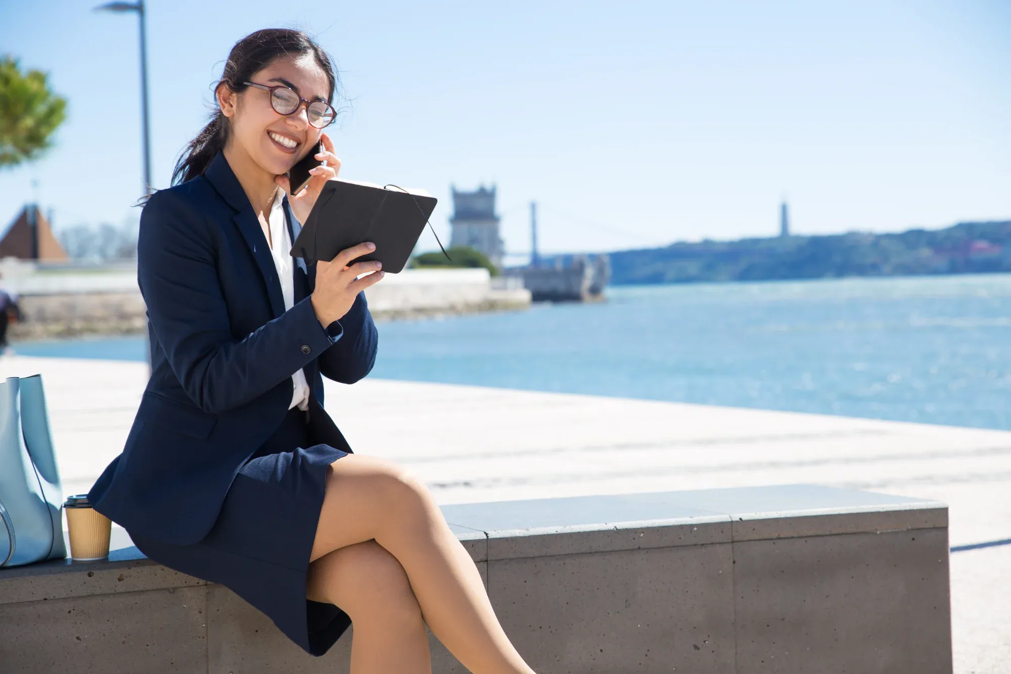 A young woman in a suit on a phone call by the water in a foreign city, tablet in hand